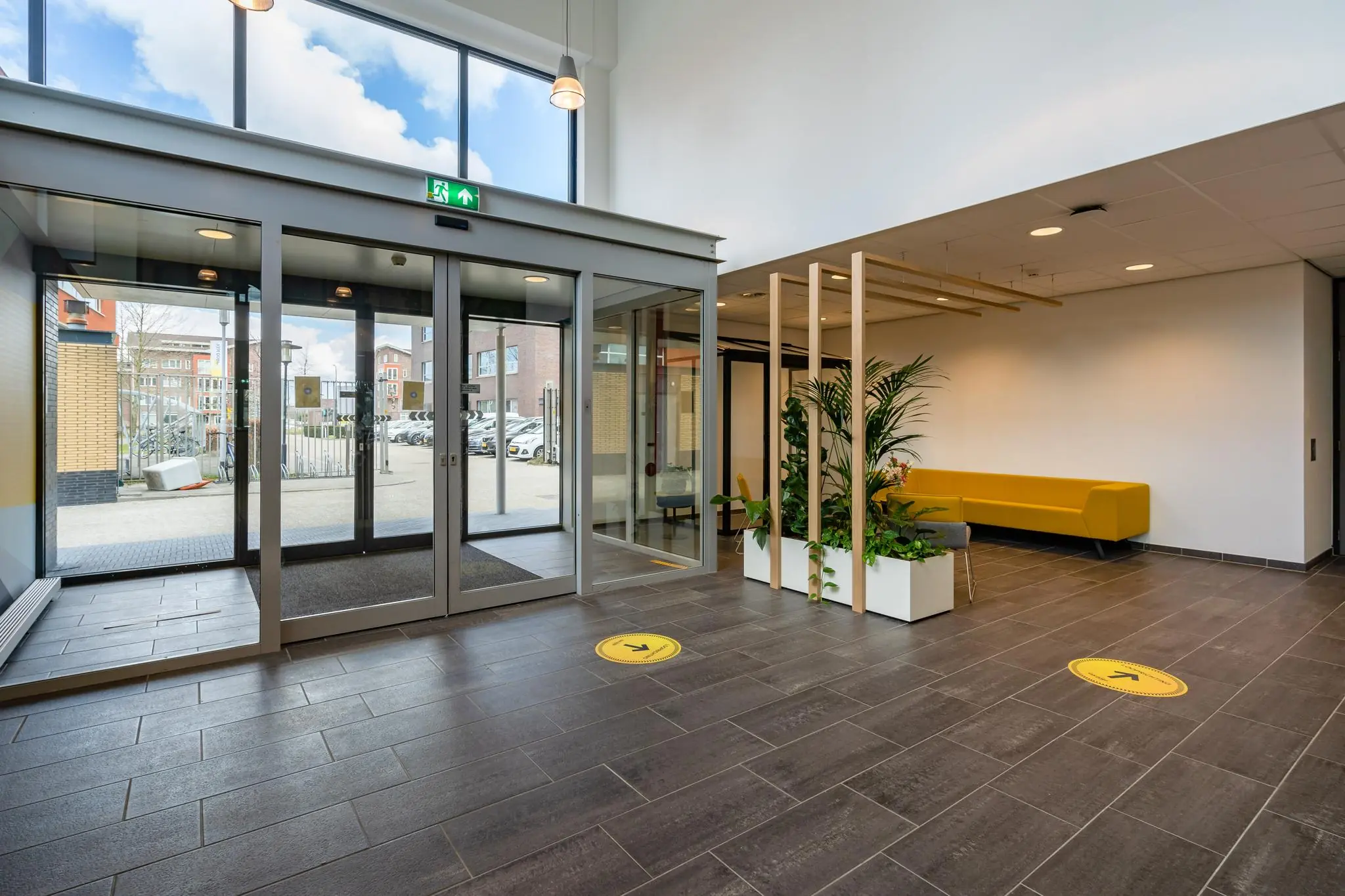 Moderne entreehal aan de Fascinatio Boulevard met glazen schuifdeuren, gele wachtbank en plantenwand.